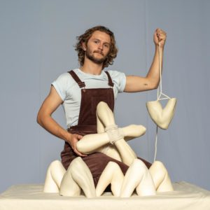 Sam dressed in brown overalls is kneeling amongst their ceramic cast elbows holding one elbow up by white cotton rope. On their lap is their sculpture "XO" made from four elbows tied together in an x shape by rope. 