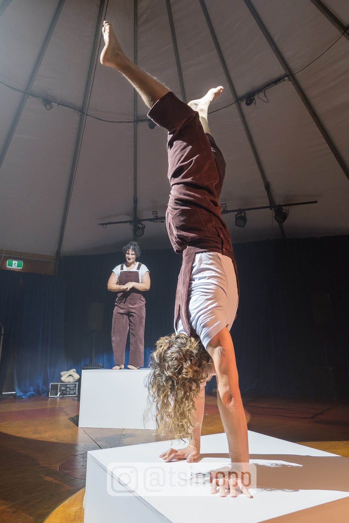 Two performers on stage atop separate large white plinths. Sam is in the foreground handstanding, Em behind presenting with adjoining arms.